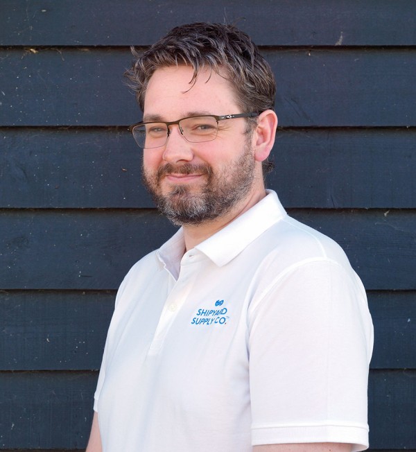 A man with brown hair in a white shirt photographed against a dark wall